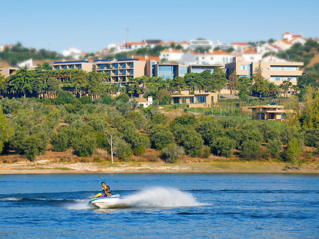 Ap Lago Montargil - Conference & Spa Экстерьер фото The photo depicts a person riding a jet ski on a body of water. In the background, there are modern buildings and lush greenery, suggesting a scenic landscape, likely near a resort or upscale residential area. The clear blue sky and calm water enhanc