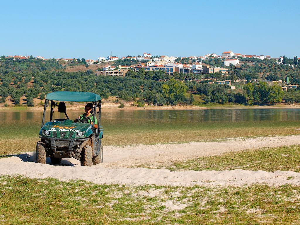 Ap Lago Montargil - Conference & Spa Экстерьер фото The photo shows a small green all-terrain vehicle driving along a sandy path beside a body of water. In the background, there's a landscape with trees and rolling hills, along with a town or village visible on the hillside. The sky is clear, indicati