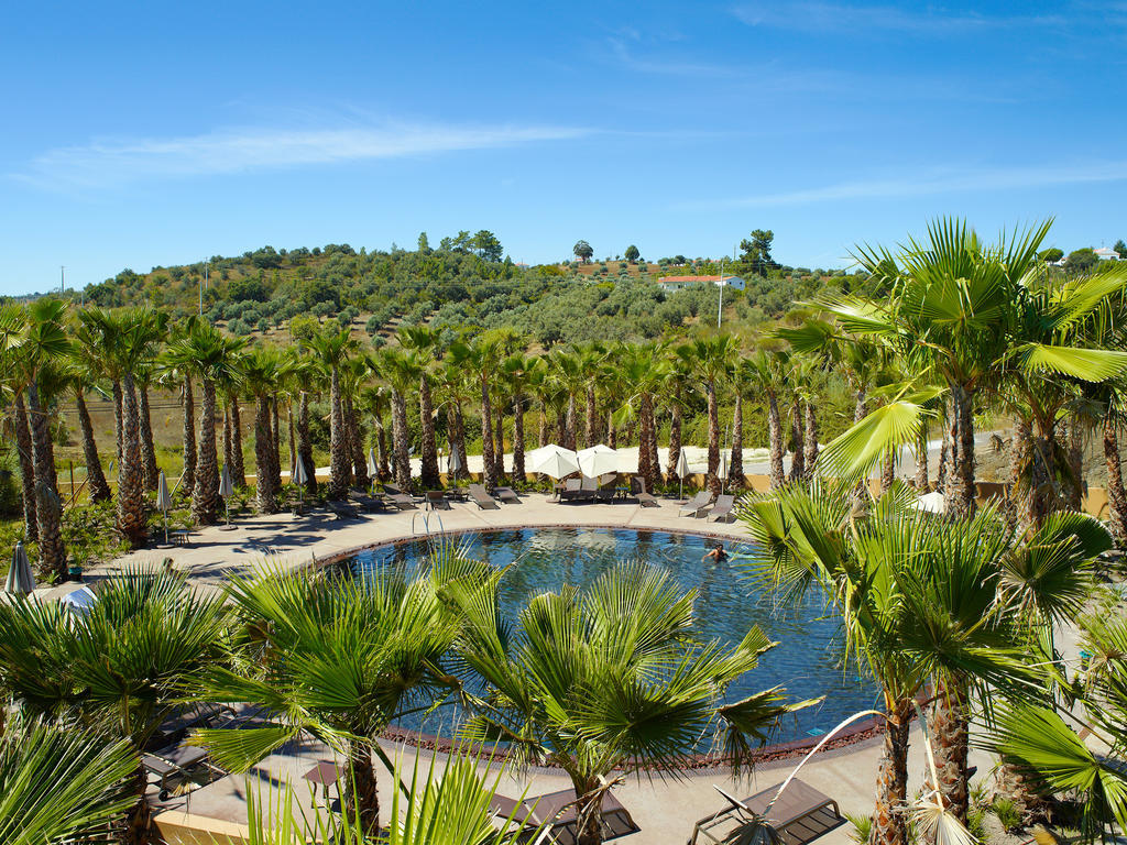 Ap Lago Montargil - Conference & Spa Экстерьер фото The photo depicts a beautiful outdoor area featuring a circular swimming pool surrounded by palm trees. The pool area has several lounge chairs arranged around it. In the background, there is a lush green hillside, and the sky is clear with a bright 