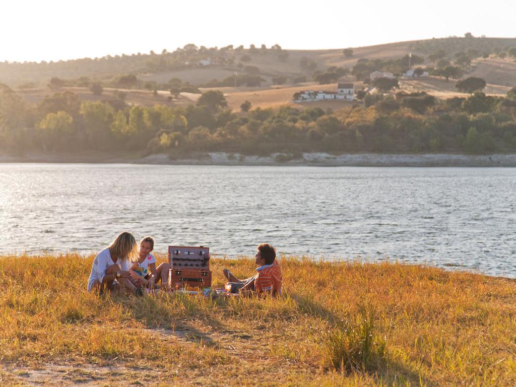 Ap Lago Montargil - Conference & Spa Экстерьер фото The photo depicts a serene lakeside scene during sunset. Three people are gathered on a grassy area near the water, enjoying a picnic. They are seated around a picnic basket, with the shimmering lake in the background and hills in the distance. The w