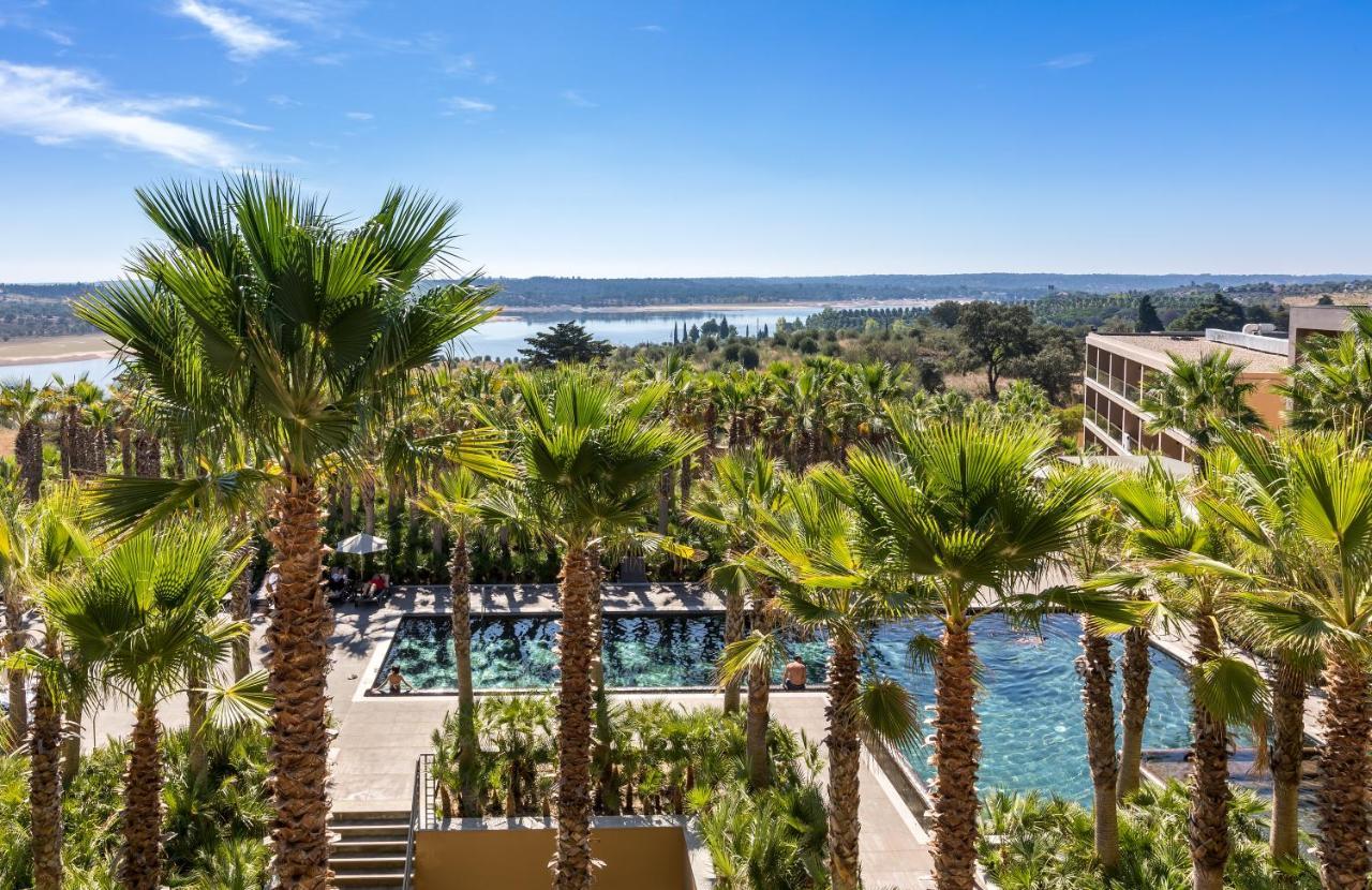 Ap Lago Montargil - Conference & Spa Экстерьер фото The photo depicts a picturesque resort landscape with a clear blue sky. In the foreground, there are tall palm trees surrounding a swimming pool area. The swimming pool has a modern design and is surrounded by lush greenery. In the background, you ca