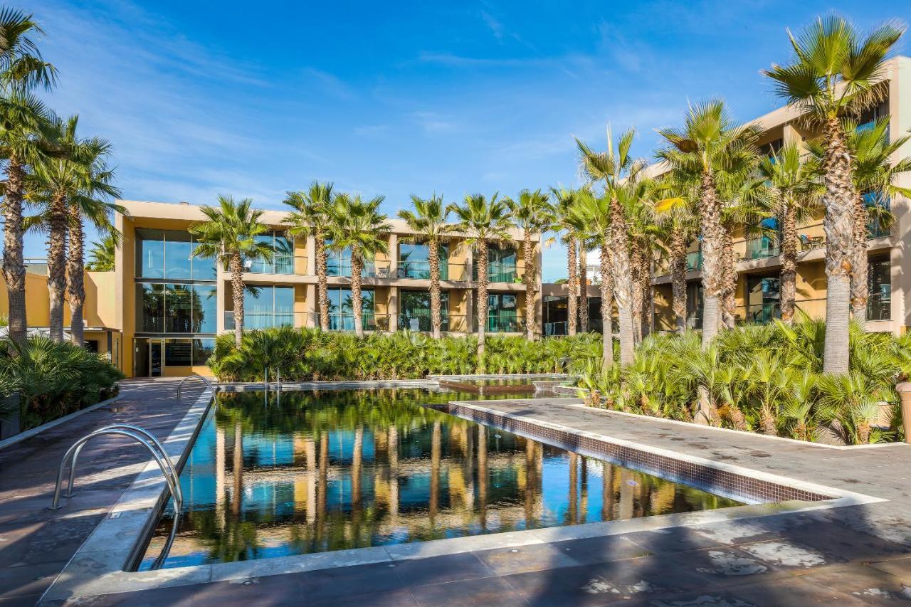 Ap Lago Montargil - Conference & Spa Экстерьер фото The photo features a modern building surrounded by lush greenery and palm trees. In the foreground, there is a serene swimming pool with a reflective surface that captures the surrounding environment. The architecture of the building appears to have 