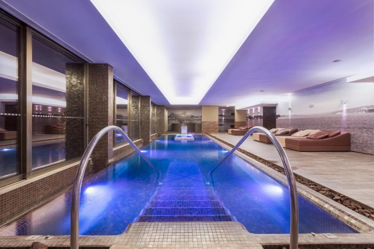 Ap Lago Montargil - Conference & Spa Экстерьер фото The photo shows a modern indoor swimming pool area. The pool features a sleek design with steps leading into the water and a gentle curve at the sides. The water is illuminated in a blue hue, enhancing the relaxing atmosphere. Surrounding the pool ar