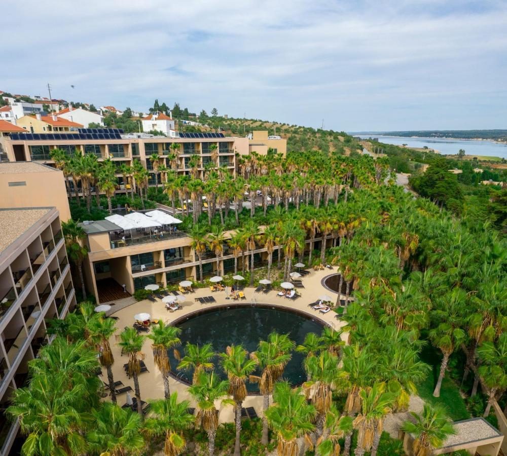 Ap Lago Montargil - Conference & Spa Экстерьер фото The photo shows a modern hotel complex set amidst lush greenery, featuring numerous palm trees. In the foreground, there is a circular swimming pool surrounded by lounge chairs. The hotel itself has multiple levels with balconies overlooking the pool