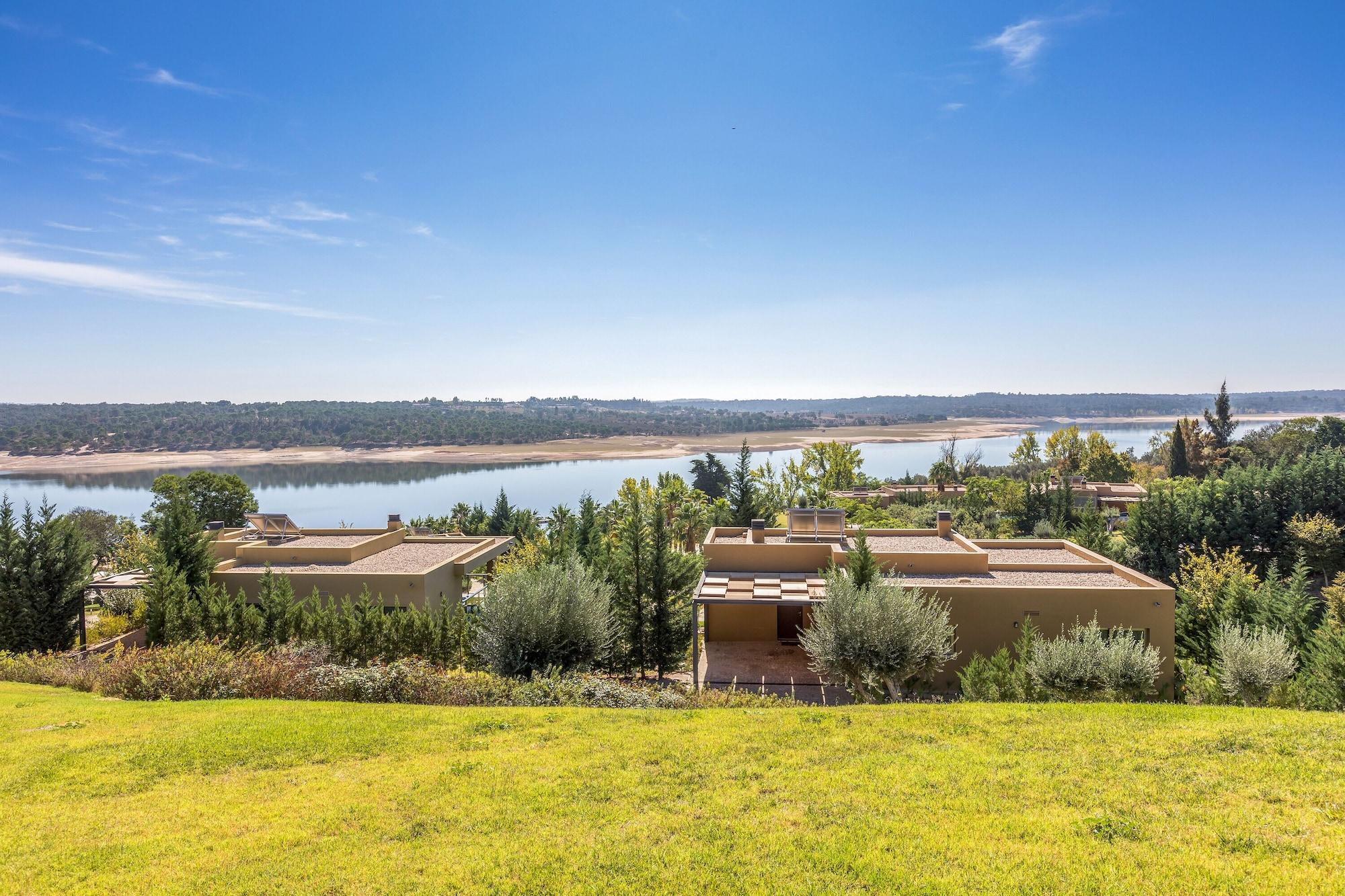 Ap Lago Montargil - Conference & Spa Экстерьер фото The photo depicts a scenic landscape featuring a calm body of water, possibly a lake or river, surrounded by lush greenery and trees. In the foreground, there are several modern buildings with flat roofs, set against a vibrant blue sky. The overall s