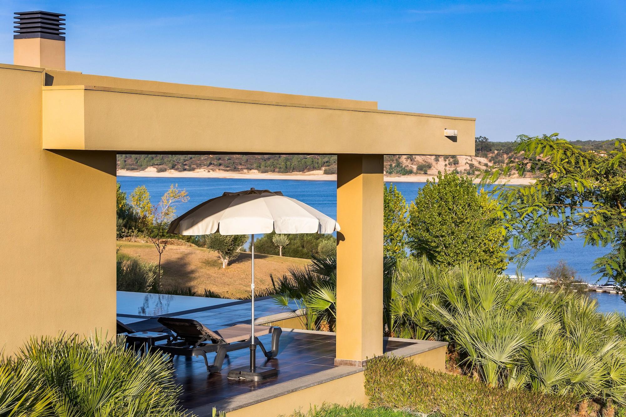 Ap Lago Montargil - Conference & Spa Экстерьер фото The photo shows a peaceful outdoor area with a modern architectural structure featuring an overhang. There is a lounge chair positioned beneath a large umbrella, creating a shaded spot to relax. In the background, there is a picturesque view of a bod