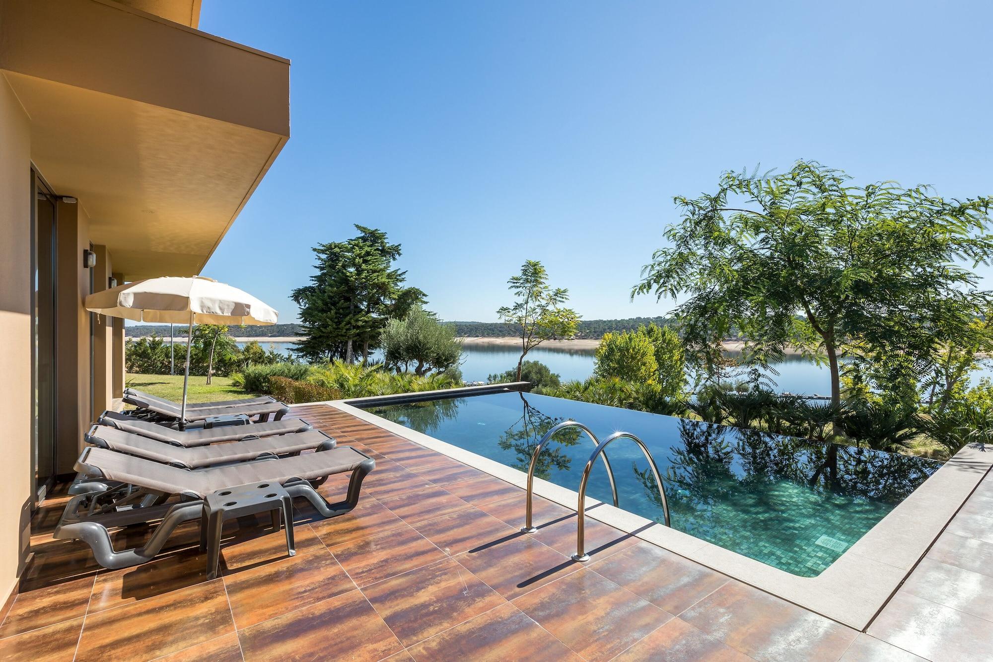 Ap Lago Montargil - Conference & Spa Экстерьер фото The photo shows a luxurious outdoor setting by a pool. There are several lounge chairs arranged next to an infinity pool, which overlooks a scenic body of water and lush greenery. A parasol provides shade for relaxation. The sky is clear and blue, in