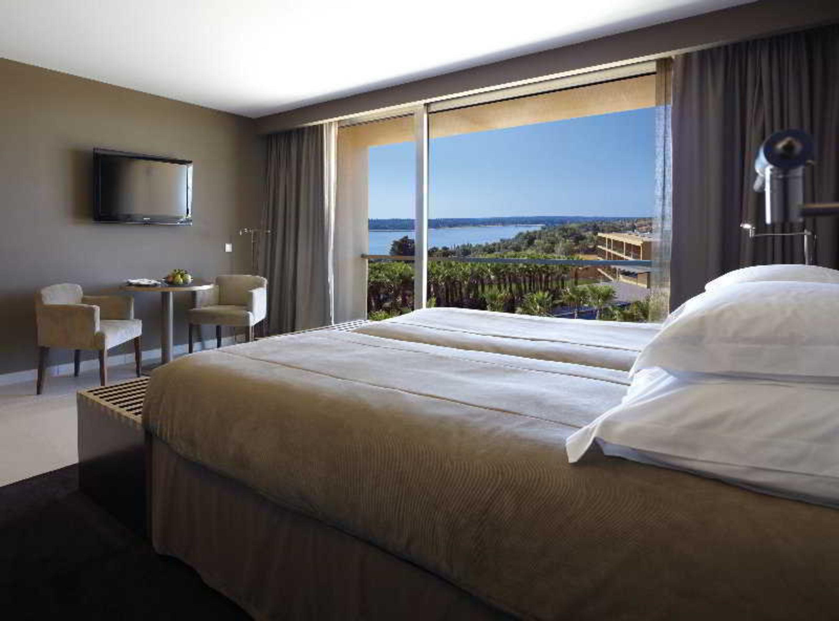 Ap Lago Montargil - Conference & Spa Экстерьер фото The photo shows a modern hotel room with a large window offering a view of the sea and mountains. The room features a comfortable bed with white pillows and brown bedding. There is a small sitting area with two chairs and a table, as well as a wall-m