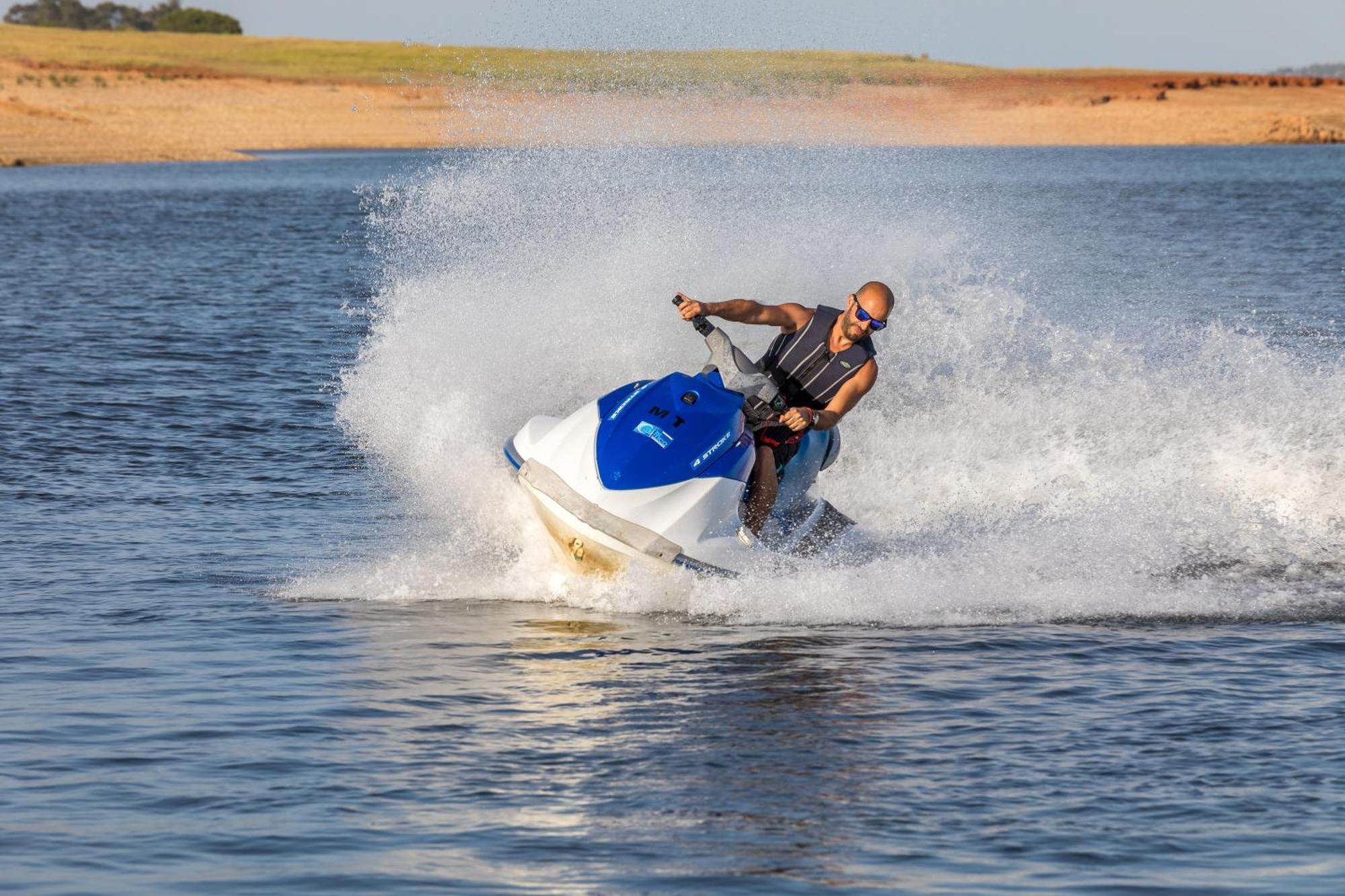 Ap Lago Montargil - Conference & Spa Экстерьер фото The photo shows a person riding a jet ski over a body of water. The rider is wearing sunglasses and a life vest, leaning forward as the jet ski accelerates, creating a spray of water around them. The background features a natural landscape, likely a 