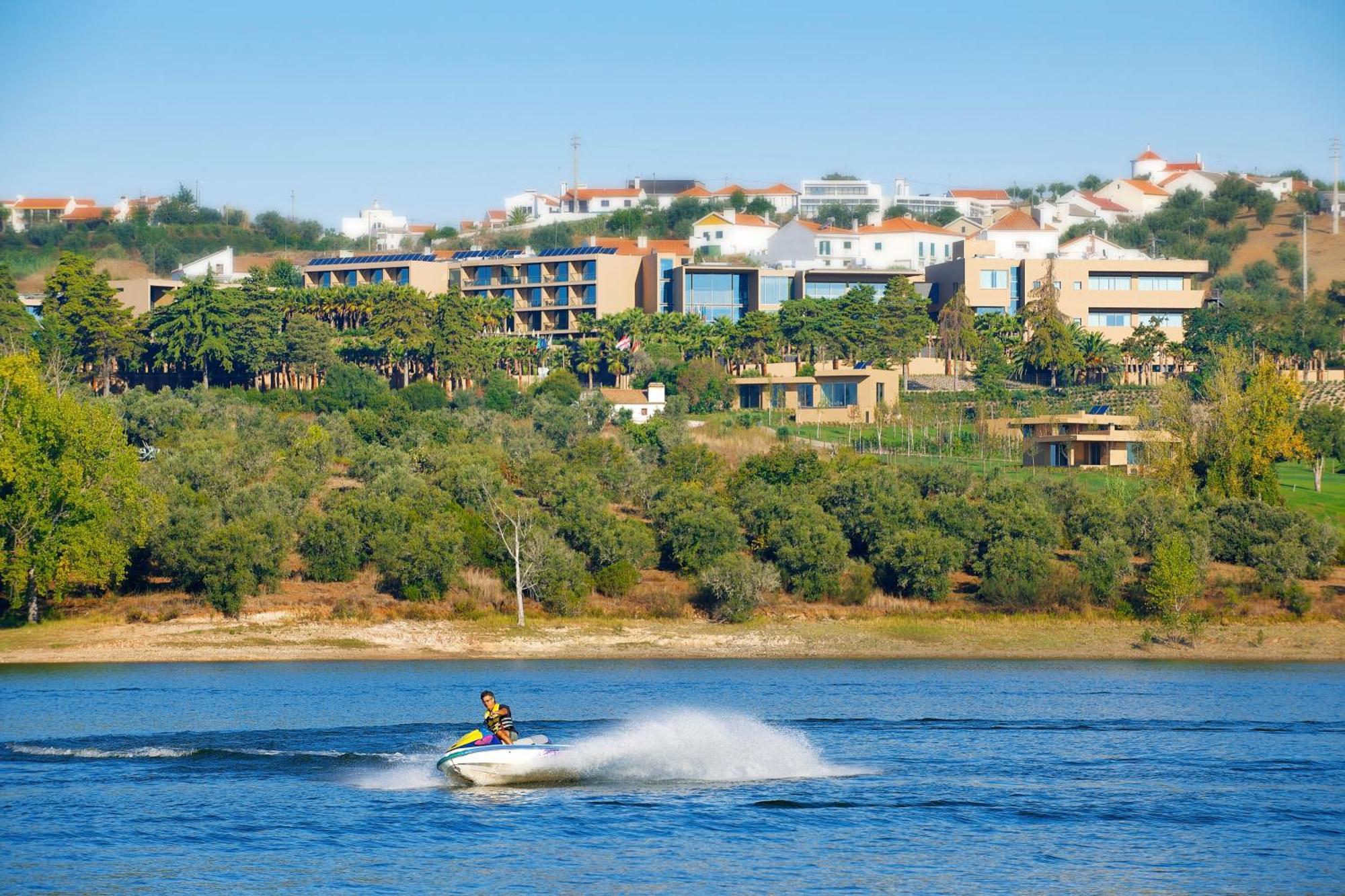 Ap Lago Montargil - Conference & Spa Экстерьер фото The photo shows a person riding a jet ski on a body of water, creating a spray as they speed across the surface. In the background, there are modern buildings and a hillside with trees and greenery. The sky is clear and blue, suggesting a bright and 
