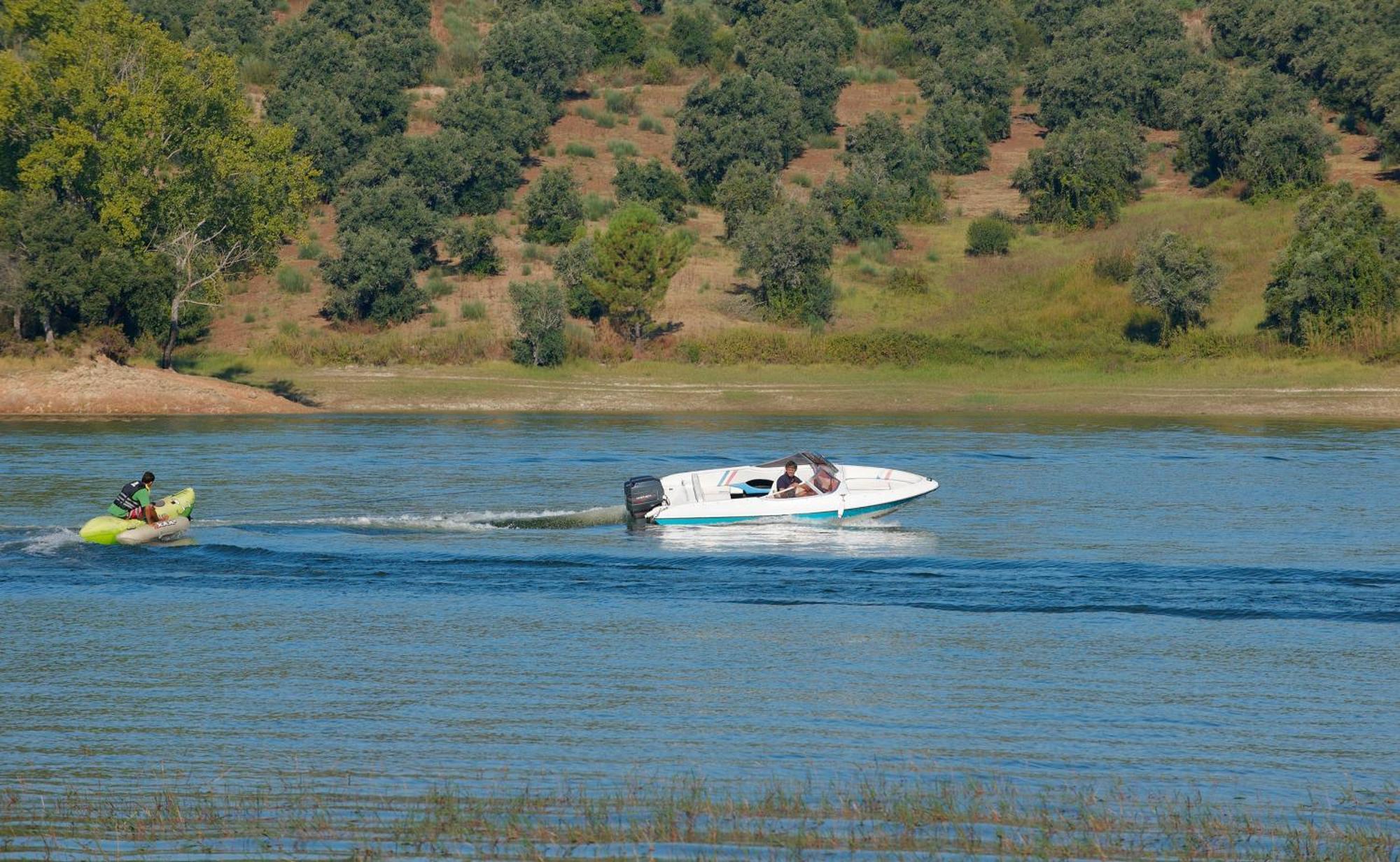 Ap Lago Montargil - Conference & Spa Экстерьер фото The photo shows a scene on a water body, likely a river or lake. In the foreground, a speedboat is moving quickly through the water, leaving a wake behind it. There are two people visible in the boat. In the background, there is a hilly area covered 