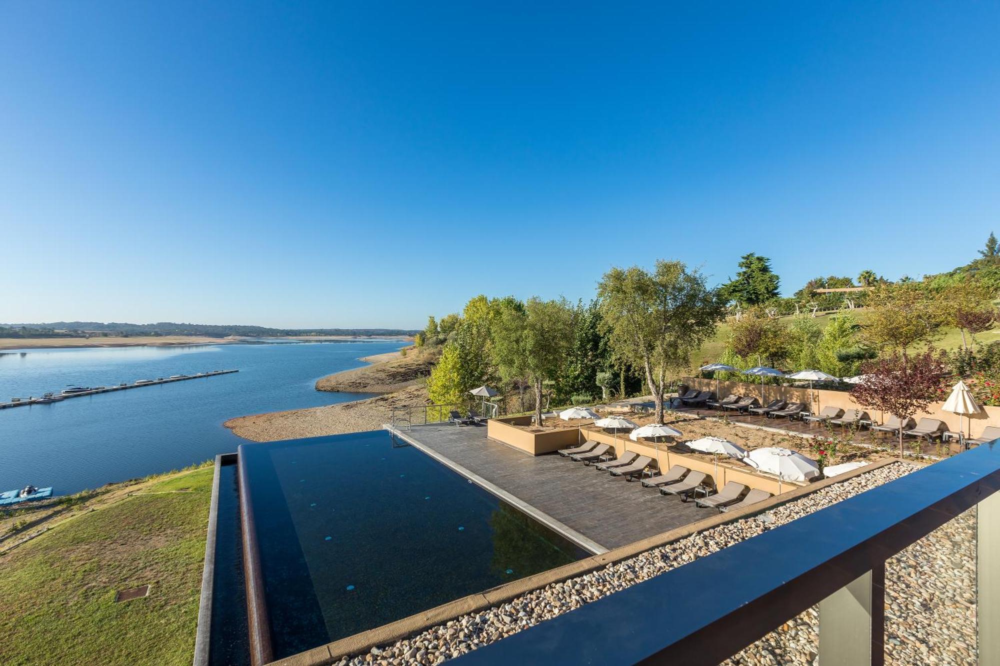 Ap Lago Montargil - Conference & Spa Экстерьер фото The photograph depicts a scenic view of a tranquil water body, likely a lake or river, bordered by lush greenery. In the foreground, there is an infinity pool that blends seamlessly with the water beyond. Comfortable lounge chairs are arranged on a w