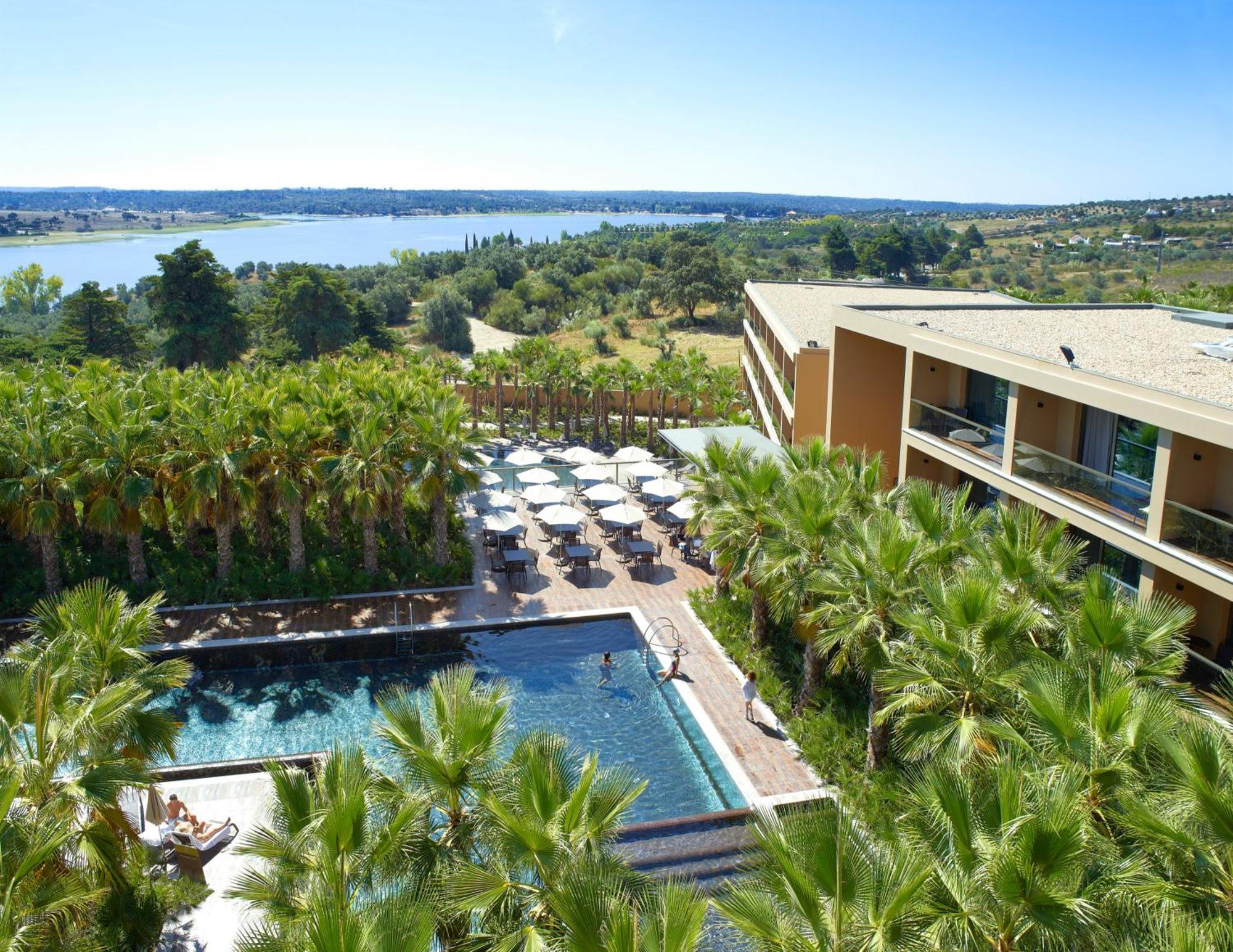 Ap Lago Montargil - Conference & Spa Экстерьер фото The photo shows a scenic view of a modern hotel or resort. In the foreground, there is a swimming pool surrounded by palm trees, with lounge chairs positioned beside it. Sun umbrellas provide shade over the chairs. The background features a lush land