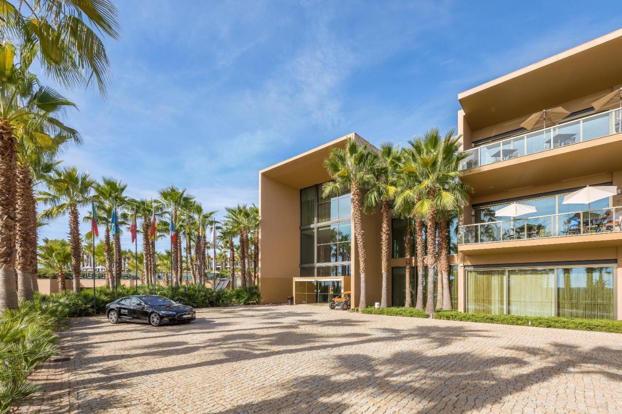 Ap Lago Montargil - Conference & Spa Экстерьер фото The photo shows a modern building with a sleek design, featuring large glass windows and balconies. The structure is surrounded by a well-maintained outdoor area with palm trees, adding a tropical feel. In front of the building, there is a paved park