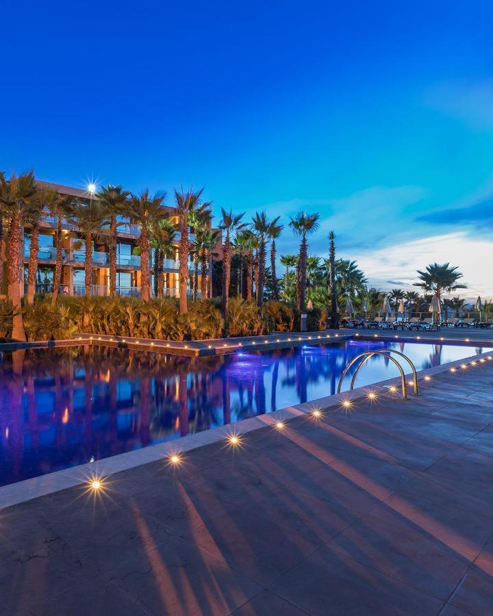 Ap Lago Montargil - Conference & Spa Экстерьер фото The image shows a serene and inviting outdoor area with a large swimming pool that features a beautiful blue hue, likely illuminated by lights. Surrounding the pool are well-maintained palm trees and lush greenery, creating a tropical atmosphere. In 